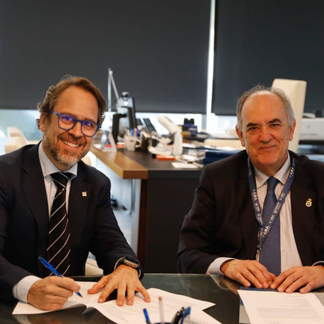 Dos hombres de traje firman documentos en una oficina moderna, sonrientes, junto a una mesa con papeles.