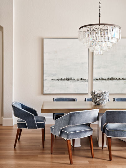 Formal dining room with sculptural dining table, blue upholstered chairs, crystal chandelier, large windows, and abundant natural light.