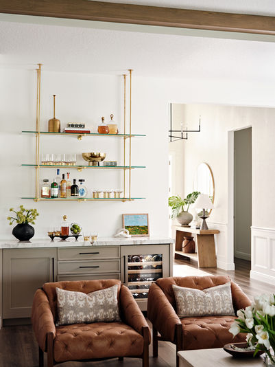 Home bar with floating glass shelves, brass hardware, built-in wine fridge, light cabinetry, and brown leather lounge chairs styled for entertaining.