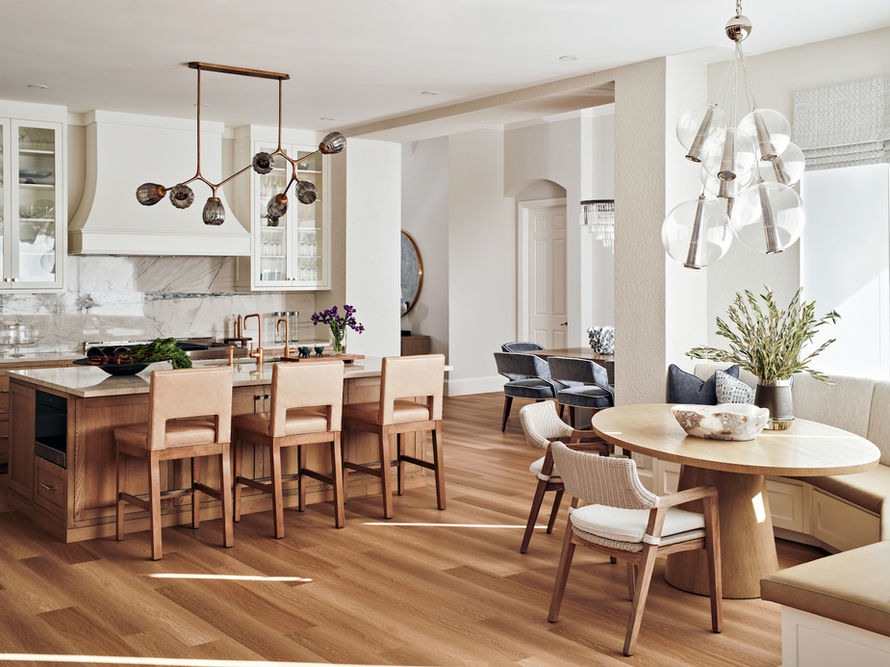 Open kitchen and dining area with neutral palette, sculptural modern chandeliers, circular wood dining table with banquette seating, and natural wood floors.