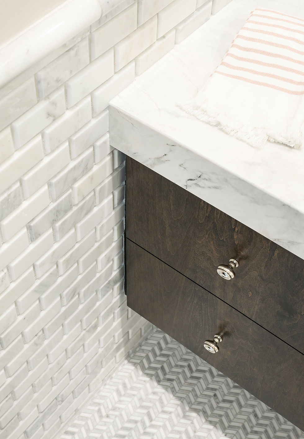 Bright bathroom corner with marble vanity, dark wood drawers, Carrera marble subway tile, Carrera chevron floor, and a striped towel on top