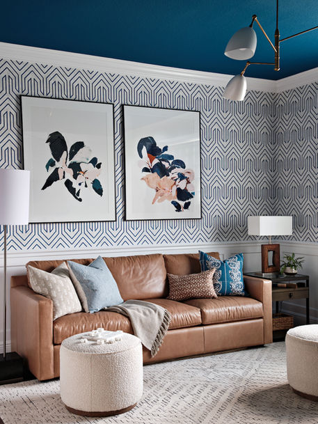 Game room with brown leather sofa, blue and neutral pillows, patterned wallpaper, framed artwork, ottomans, and modern ceiling light.
