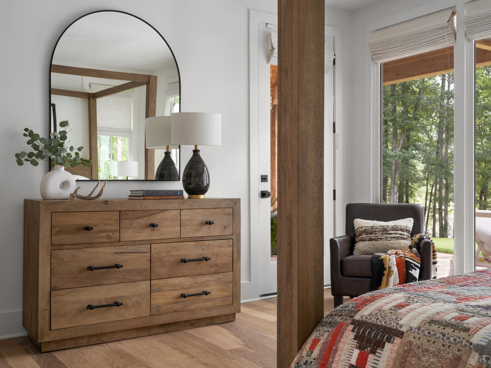 Modern bedroom with a wooden dresser, round mirror, books, table lamps, and an armchair near large windows overlooking a forest.
Palmer Kay Design_Interior Design_Peachtree City, GA