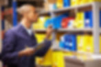 Man in blue overalls checks inventory on metal shelves with yellow, blue, and red bins, holding a clipboard and pencil, in warehouse.