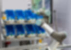 Robotic arm moves between blue bins on a metal rack in an industrial setting. The scene is bright and organized, with a focused mood.