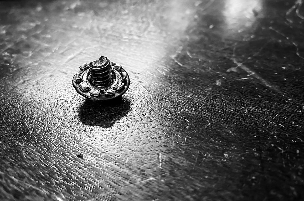 Close-up of a single screw on a textured, dark surface. The screw casts a shadow, with a metallic, reflective appearance. Black and white.