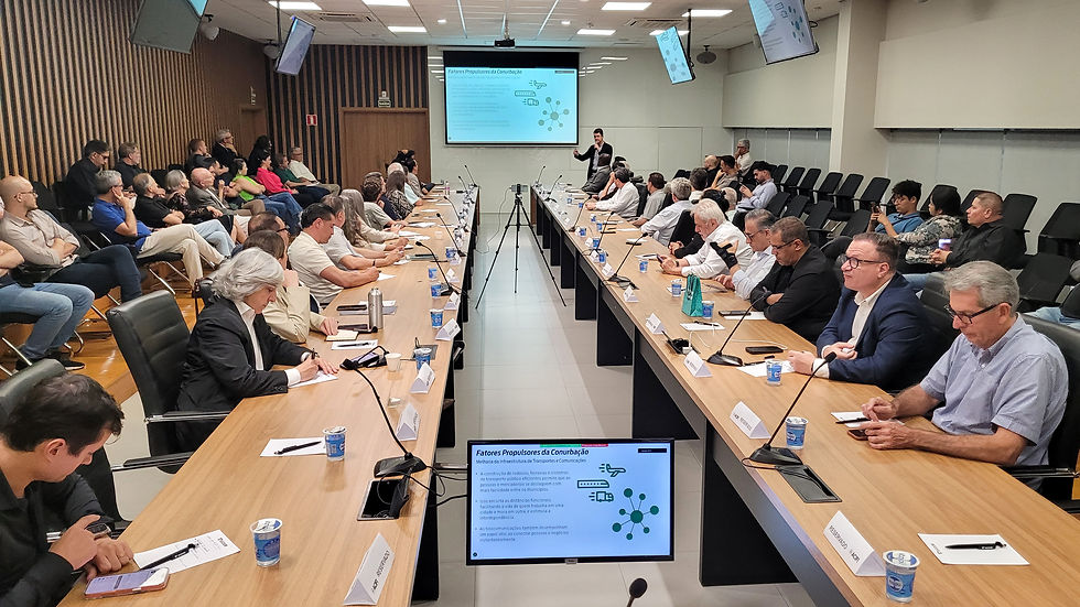 Fotografia de uma reunião ampla realizada em uma sala de conferências, com duas longas mesas retangulares ocupadas por participantes dos dois lados. Várias pessoas estão sentadas tomando notas, enquanto uma pessoa apresenta slides projetados na tela frontal. Monitores adicionais mostram o conteúdo da apresentação. Há copos, papéis e placas de identificação sobre a mesa.