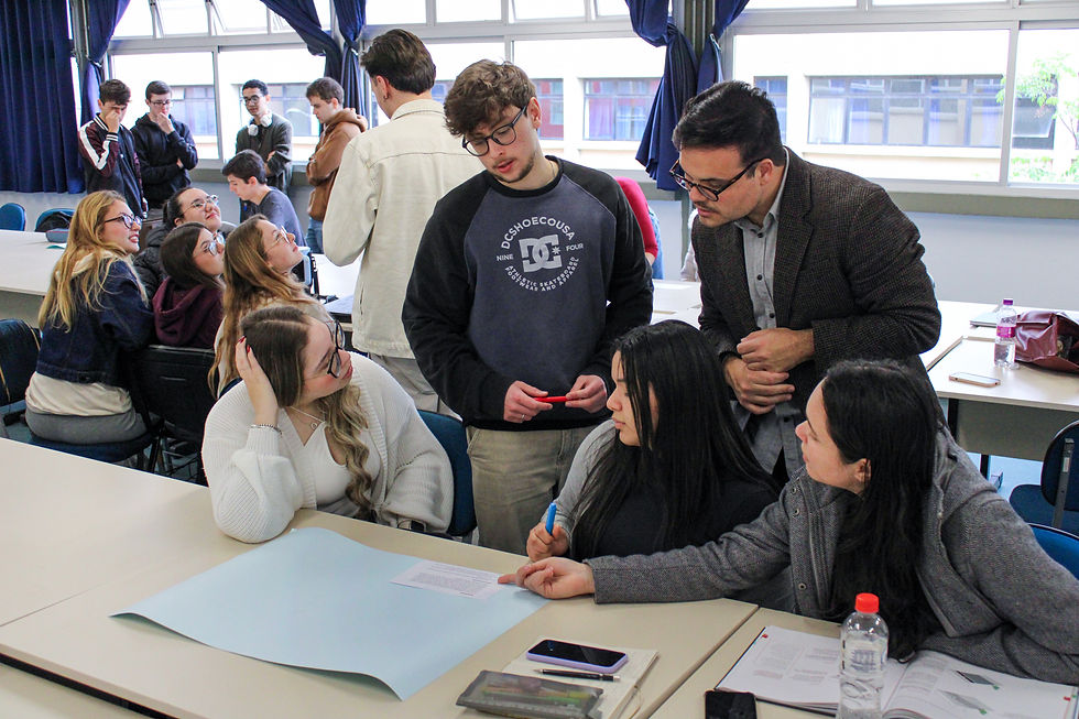 Fotografia de um grupo reunido em torno de uma mesa, dialogando e escrevendo em uma folha grande de papel azul. Outra pessoa se inclina para acompanhar a discussão, enquanto o restante da turma aparece ao fundo.