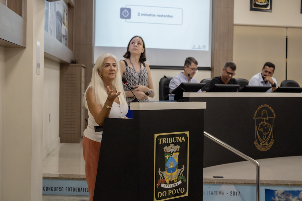 Fotografia de uma mulher de cabelos brancos e blusa branca discursando na tribuna de um plenário. Ao fundo, uma mulher e três homens acompanham o discurso.