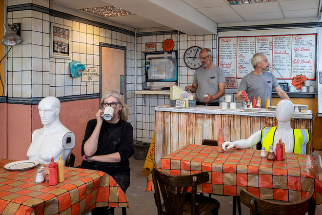 the York based Bombsquad Art collective pose for a team shot in a cafe created from cardboard by the artist Jo Peel. One woman is sat down drinking tea next to a mannequin while two men are stood behind the counter.