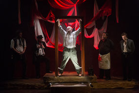 a male actor is suspended in a wooden frame on stage flanked either side by four actors largely in shadow. He is in obvious pain, accentuated by the red stage lighting