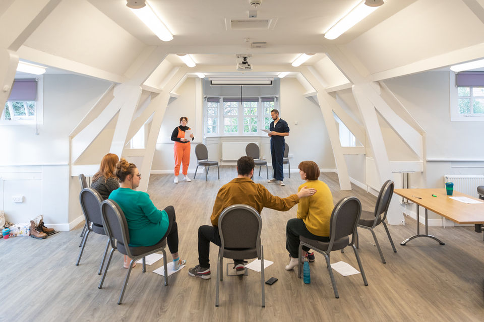 theatre - actors rehearsing for a play in a white room. Four actors are sat in a semi circle while two actors are standing reading lines