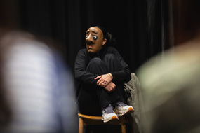 an actor dressed in black wearing a full mask with a startled expression sitting on a chair