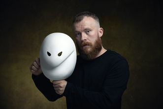an actor wearing black holds a larval mask with the expression 'trouble'