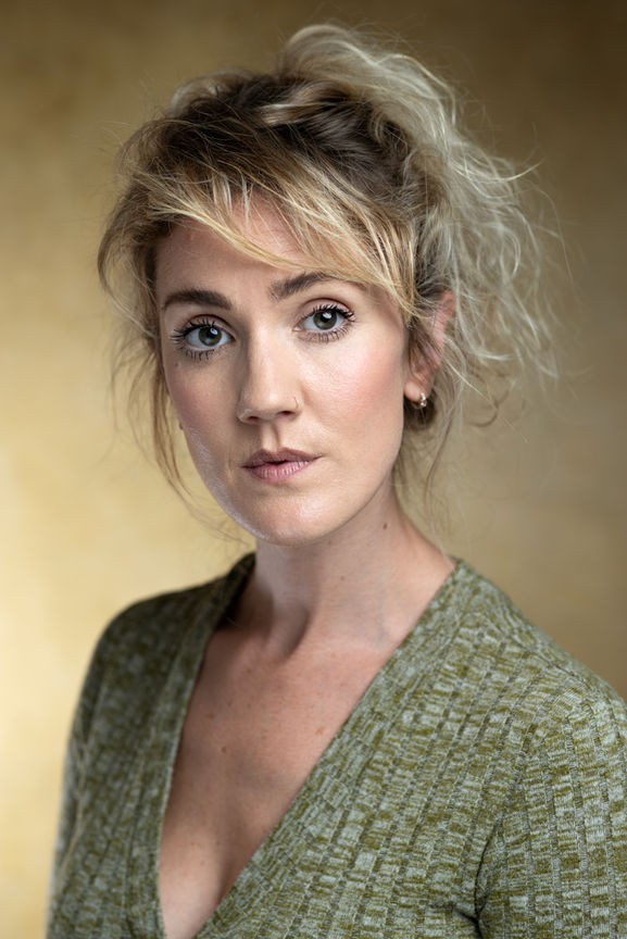 headshots - a female actor with fair hair and wearing a light green top poses against a beige backdrop