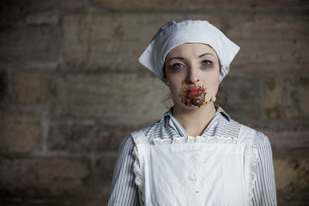 A young female actor in a Nurse uniform stands in front of a stone interior wall. Her face is smeared with chocolate.