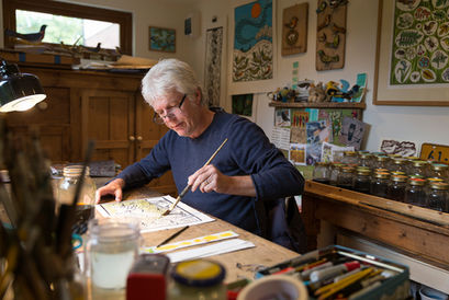 a seated artist at work in his York home studio. he is holding a paint brush, surrounded by paint and bright lino prints