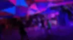People dance under vibrant, colorful tent at night. DJ performs on stage. Energetic, lively atmosphere with purple and blue lighting.