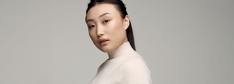 Sophie is an asian woman, wearing a white blouse. She has her hair up, and is standing in front of a beige wall.
