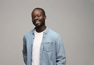 Elijah is a Ghanian man, wearing a tensil blue shirt with a white tee. He is standing against a beige background.