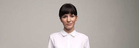 Olivia is a mixed race woman with dark hair. She is wearing a white oxford and is standing against a beige background.
