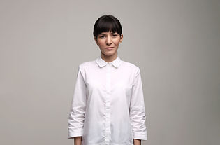 Olivia is a mixed race woman with dark hair. She is wearing a white oxford and is standing against a beige background.