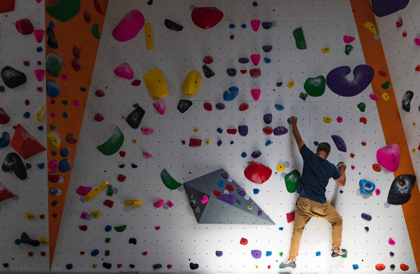 Man climbing a colorful indoor rock wall, wearing a blue shirt and tan pants. The wall features bright holds and an orange panel.