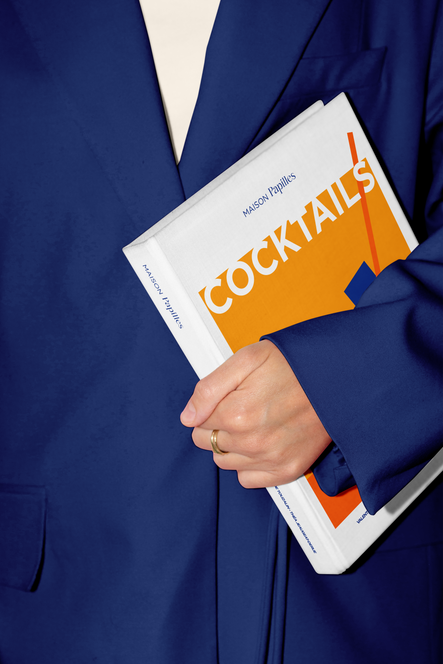 Personne tenant livre de recette de cocktails, couverture orange sur fond bleu foncé, projet de mise en page et analyse marketing, design graphique, direction artistique par Valentine carré