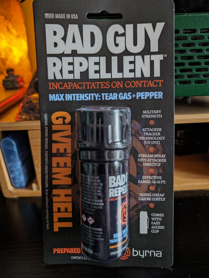 "Bad Guy Repellent" spray on black packaging with bold text. Promises incapacitation using tear gas and pepper. Shelf and orange light in background.