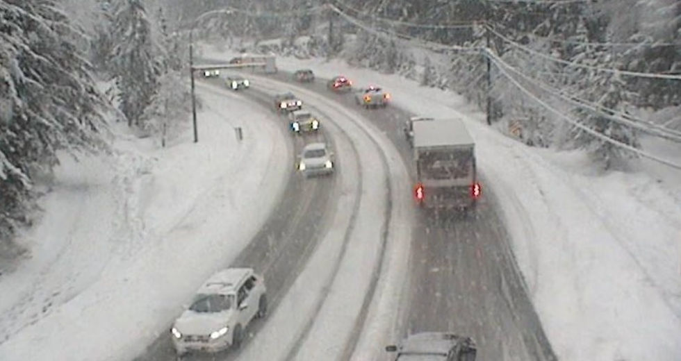 Snow‑covered section of the Sea to Sky Highway near Squamish with an Environment Canada snowfall warning graphic displayed, indicating hazardous winter driving conditions.