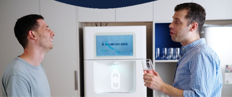 Two men chatting near a water dispenser, one holds a glass. Background shows cabinets and a digital display. Mood appears friendly.