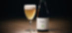 A bottle and glass of Sofie beer on a wooden table. The glass is filled with light golden beer topped with foam. Dark, simple background.