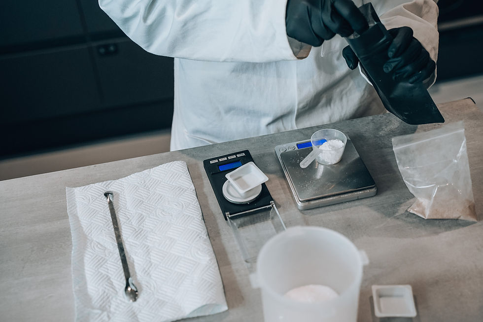 Gloved person in a lab coat measures white powder on digital scales beside a spoon and plastic bag on a stainless counter.