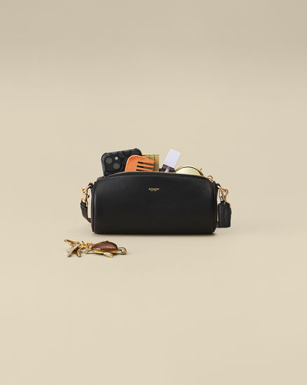 Black Barrel Coach bag unzipped to reveal a phone, wallet, cardholder, and small items, with keys placed in front on a neutral background