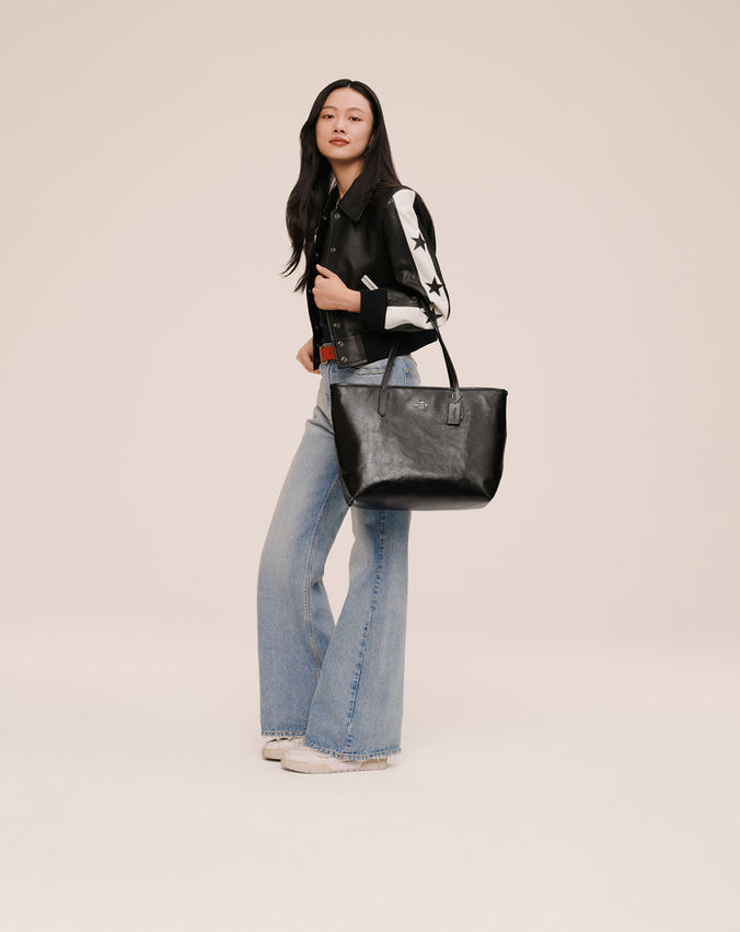 Model in a black leather jacket and wide-leg jeans carries a black Coach city tote with long handles against a neutral cream background.