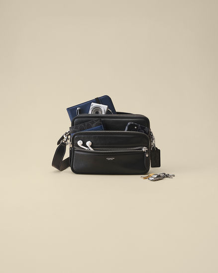 Black Dakota Coach crossbody bag with multiple zip compartments, open to display a camera, wallet, earbuds, phone, and keys on a neutral background.