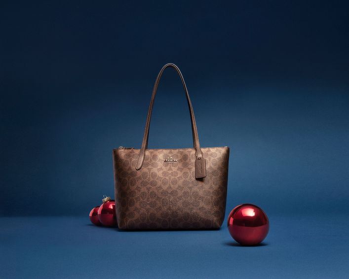 Fiona Zip Tote in Metallic Signature Canvas in Brown Copper displayed with red holiday ornaments against a deep blue background.