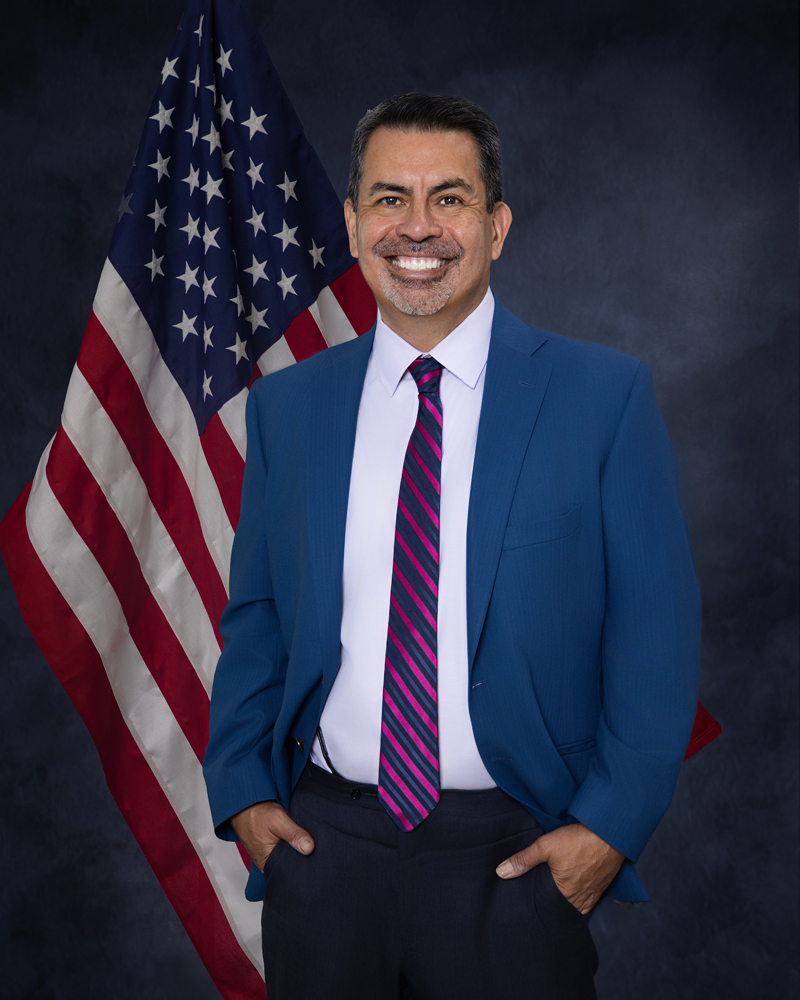 Professional headshot of a man in a suit and tie standing in front of an American flag, by Tina Dwyer Photography El Paso
