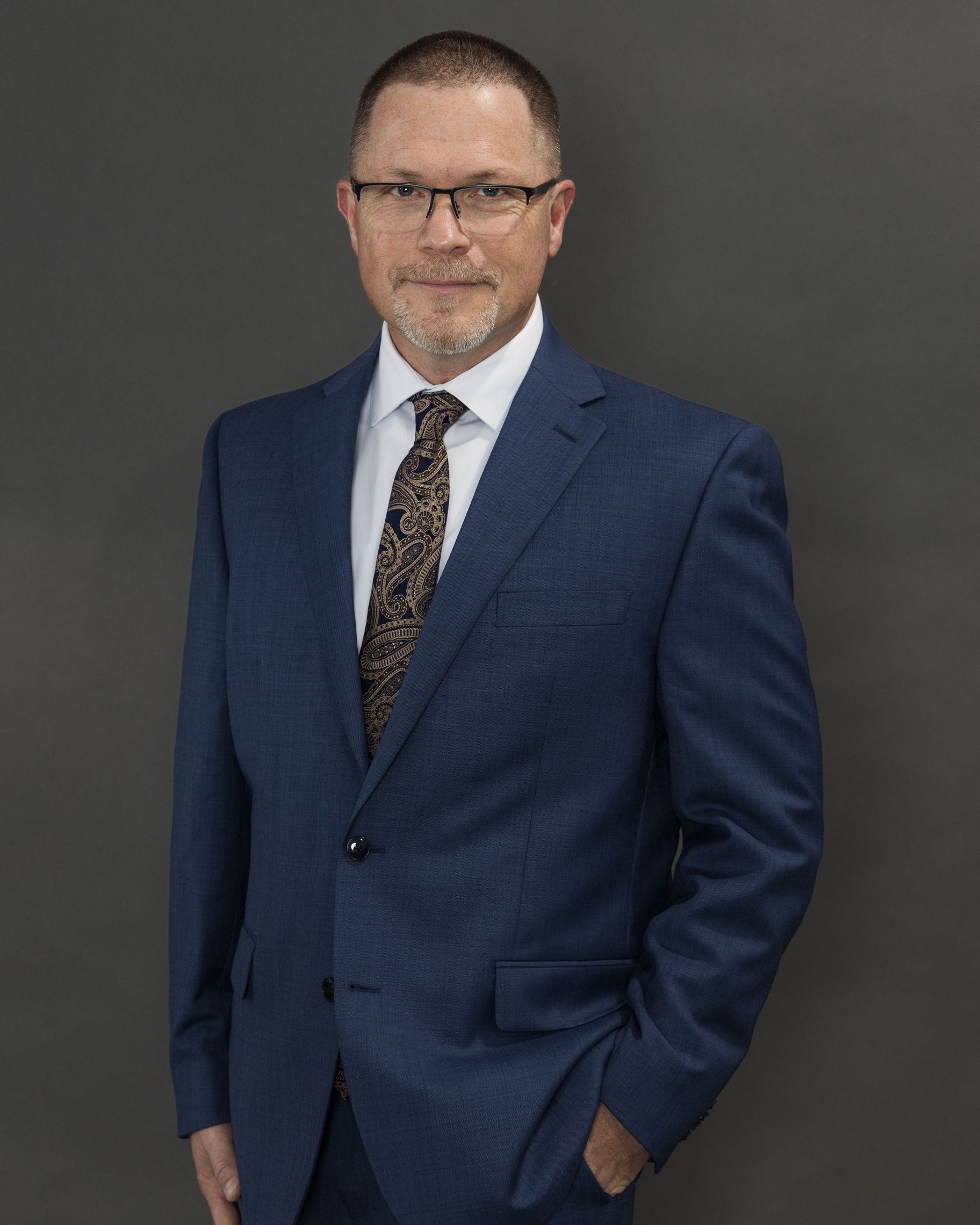 Professional headshot of a man in a dark blue suit and patterned tie against a dark background, by Tina Dwyer Photography