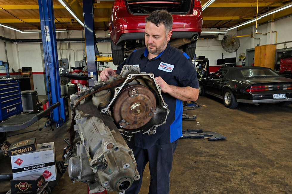 Mechanic examines a car part in a garage. Red car on lift; black car in background. Boxes labeled "PENRITE" and "MULTI-VEHICLE" nearby.