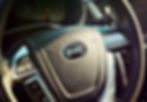 Close-up of a car steering wheel featuring the BYD logo. Black interior with control buttons visible. Sunlight highlights the textures.