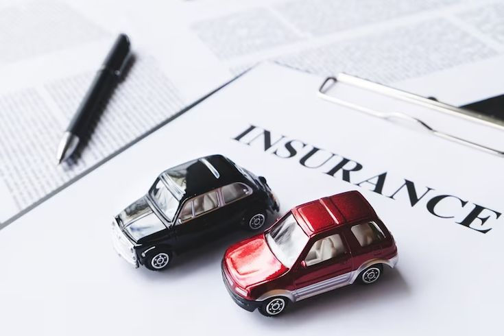 Two toy cars, one black and one red, on a document labeled "INSURANCE." A pen and clipboard are nearby, suggesting an office setting.