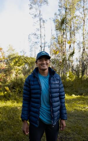 Smiling man wearing a blue jacket and hat, standing in nature setting.