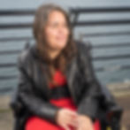 A woman in a black jacket and red dress, in a wheelchair, with water behind her