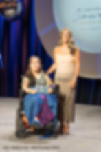 A woman in a blue dress, in a wheelchair, holding an award. A woman in a beige  dress is standing next to her.