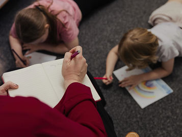 Two children lying on the floor, drawing with pencils while an adult observes and takes notes, assessing their development or behaviour.