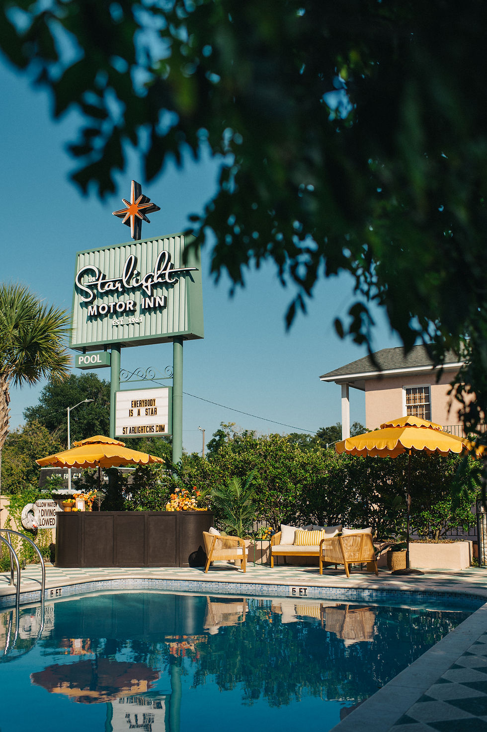 Poolside view at Starlight Motor Inn, featuring a retro sign, yellow umbrellas, wicker chairs, and flowers from The French Eclectic event rentals. Sign reads "Everybody is a star."
