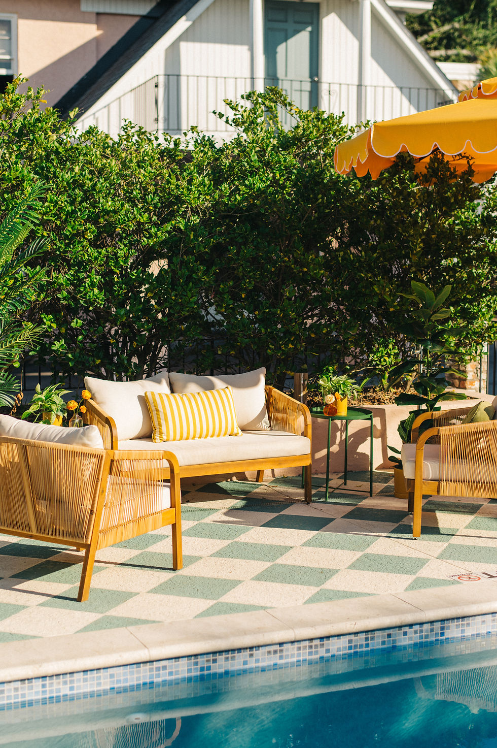 Poolside seating area with wooden chairs and a yellow-striped cushion, set on a checkered patio surrounded by lush greenery and a yellow umbrella. Private event furniture rentals in Charleston, SC