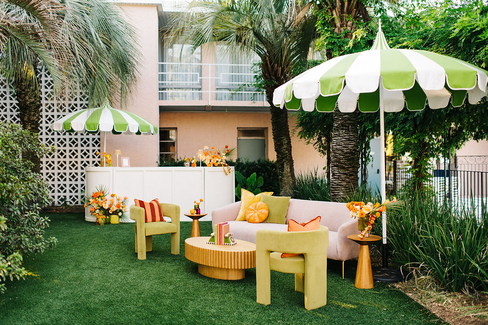 Outdoor seating area with green and white umbrellas, pink sofa, green chairs, and orange accents. Lush greenery and flowers surround.