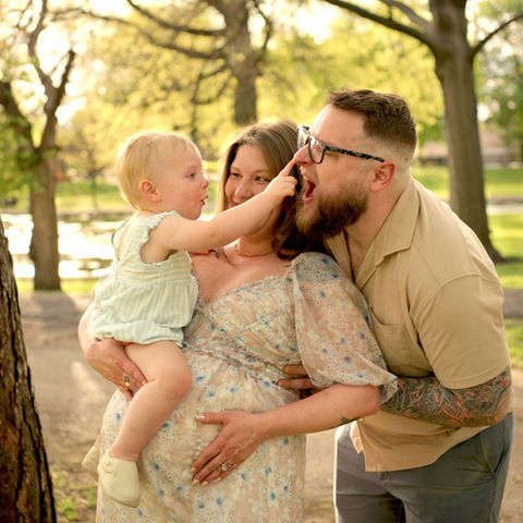 Pittsburgh family playfully interacts at Allegheny Commons Park. The pregnant mother is holding her toddler while the father laughs.
